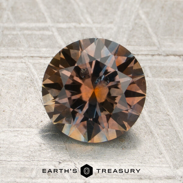 1.94-Carat Orange-Pale Blue Particolored Montana Sapphire (Heated) A 1.94-carat orange-pale blue particolored Montana sapphire (heated), cut in a round shape, rests on a textured surface with the text “EARTH’S TREASURY” displayed below the stone.