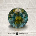 A 1.81-carat teal blue-golden yellow particolored Montana sapphire, round and faceted, sits on a textured surface. “Earth’s Treasury” is written at the bottom of the image.