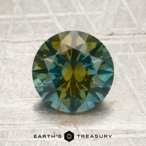 A 1.81-carat teal blue-golden yellow particolored Montana sapphire, round and faceted, sits on a textured surface. “Earth’s Treasury” is written at the bottom of the image.