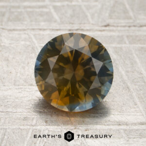1.78-carat Sky Blue-Orange Particolored Montana Sapphire (Heated) in a round faceted cut, showcasing a vibrant mix of yellow, brown, and blue hues, displayed on a textured surface with the Earth’s Treasury logo visible below.