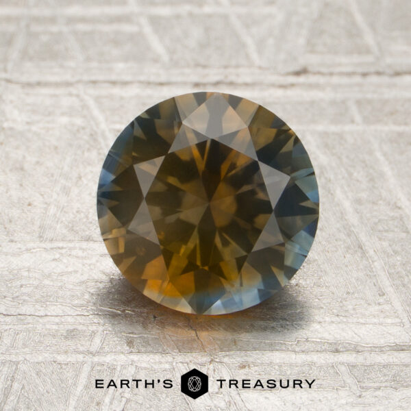 1.78-Carat Sky Blue-Brownish Orange Particolored Montana Sapphire (Heated) 1.78-carat Sky Blue-Orange Particolored Montana Sapphire (Heated) in a round faceted cut, showcasing a vibrant mix of yellow, brown, and blue hues, displayed on a textured surface with the Earth’s Treasury logo visible below.