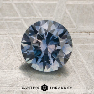 A 1.76-carat medium blue-lilac particolored Montana sapphire (heated), expertly cut in a round brilliant shape with striking geometric facets, is displayed on a textured gray surface. The Earth’s Treasury logo appears at the bottom.