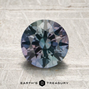 A 1.75-carat Montana sapphire (heated), cut in a round brilliant style and showing blue-green and purple hues, displayed on a textured light surface; “EARTH’S TREASURY” text and logo at the bottom.