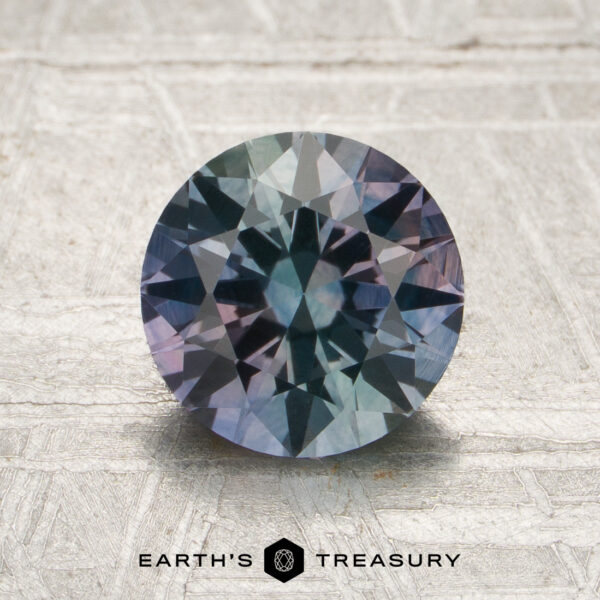1.75-Carat Blue-Purple-Green Particolored Montana Sapphire (Heated) A 1.75-carat Montana sapphire (heated), cut in a round brilliant style and showing blue-green and purple hues, displayed on a textured light surface; “EARTH’S TREASURY” text and logo at the bottom.