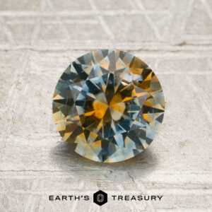 1.61-carat sky blue-orange particolored Montana sapphire with round faceting and vibrant blue and yellow color zoning, displayed on a textured surface with the “Earth’s Treasury” branding below.