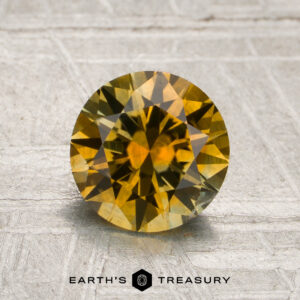 A round, faceted 1.56-carat golden yellow Montana sapphire (heated) is displayed on a textured, light-colored surface with the text “Earth’s Treasury” below.