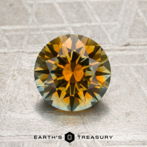 A 1.55-carat orange and pale blue particolored Montana sapphire (heated) with a round brilliant cut is displayed on a textured surface, featuring the “Earth’s Treasury” logo and text at the bottom.