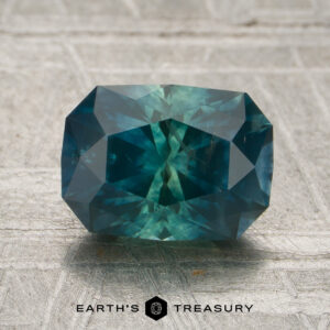 A rectangular-cut 1.91-carat Montana sapphire (heated) in a vibrant teal hue is displayed on a textured gray surface with the text “EARTH’S TREASURY” beneath it.
