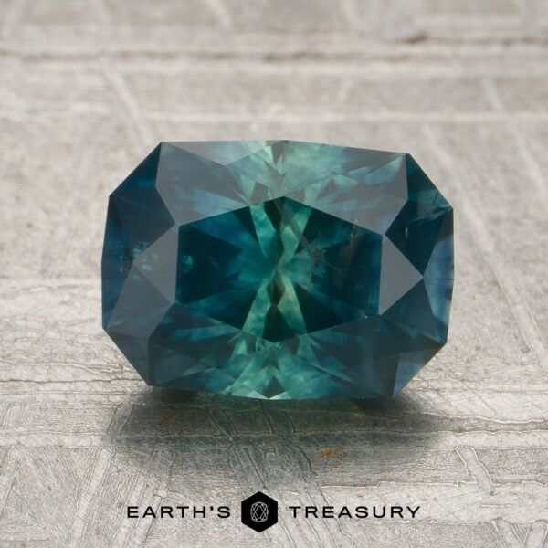 1.91-Carat Green-Teal Particolored Montana Sapphire (Heated) A rectangular-cut 1.91-carat Montana sapphire (heated) in a vibrant teal hue is displayed on a textured gray surface with the text “EARTH’S TREASURY” beneath it.