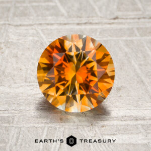 1.54-carat Vivid Orange-Colorless Particolored Montana Sapphire, round-cut, displayed on a textured surface with “Earth’s Treasury” written beneath.