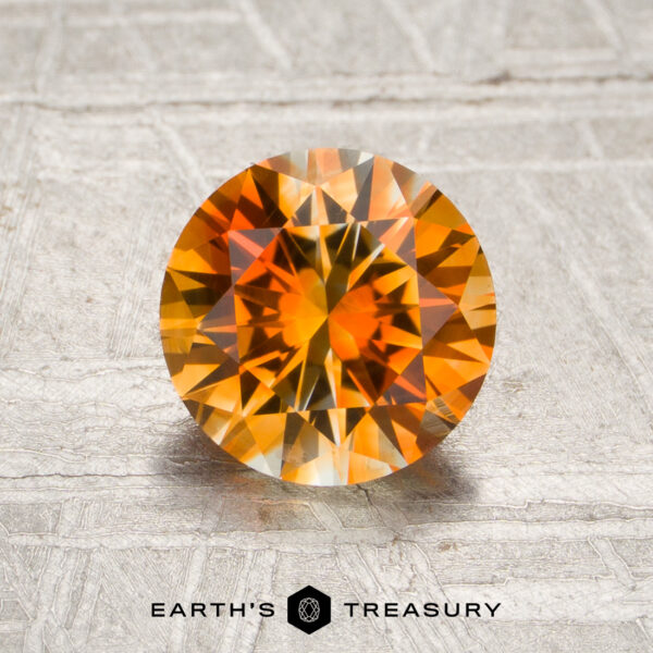 1.54-Carat Vivid Orange-Colorless Particolored Montana Sapphire (Heated) 1.54-carat Vivid Orange-Colorless Particolored Montana Sapphire, round-cut, displayed on a textured surface with “Earth’s Treasury” written beneath.