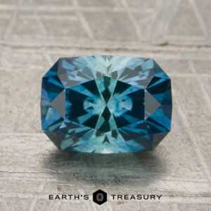 A 1.81-carat Montana sapphire (heated) with a radiant cut and starburst pattern is displayed on a textured gray surface above the words “Earth’s Treasury.”.