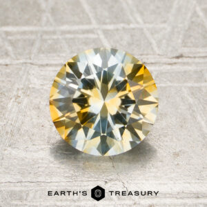1.53-carat colorless-yellow particolored Montana sapphire (heated) in a round brilliant cut, featuring yellow and blue hues, displayed on a textured metallic surface with the “Earth’s Treasury” logo and text at the bottom.