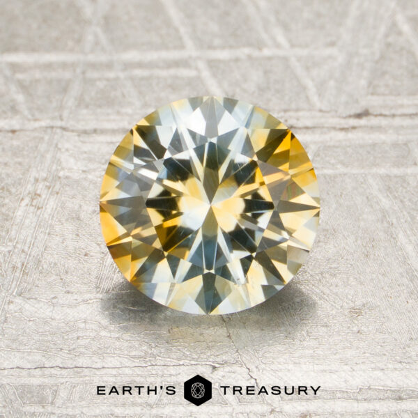 1.53-Carat Colorless-Yellow Particolored Montana Sapphire (Heated) 1.53-carat colorless-yellow particolored Montana sapphire (heated) in a round brilliant cut, featuring yellow and blue hues, displayed on a textured metallic surface with the “Earth’s Treasury” logo and text at the bottom.