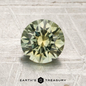 A round, faceted 1.50-carat mint green Montana sapphire (heated) with greenish-yellow hues is displayed on a textured light surface, featuring the “Earth’s Treasury” logo and text at the bottom.