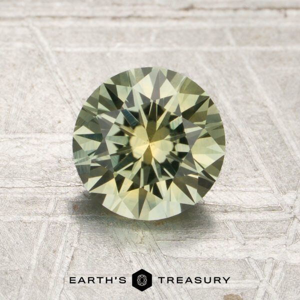 1.50-Carat Mint Green Montana Sapphire (Heated) A round, faceted 1.50-carat mint green Montana sapphire (heated) with greenish-yellow hues is displayed on a textured light surface, featuring the “Earth’s Treasury” logo and text at the bottom.