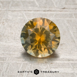 A 1.43-carat opalescent golden yellow Montana sapphire (heated), cut in a round brilliant style, shimmers with yellow and orange hues as it reflects light on a textured surface; the “Earth’s Treasury” logo is featured at the bottom of the image.