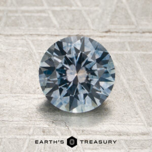 A 1.43-carat steely sky blue Montana sapphire (heated) with a round brilliant cut is displayed on a light textured surface. The Earth’s Treasury logo appears at the bottom.
