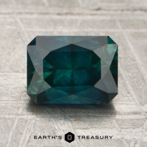 A rectangular, deep blue-green 1.89-carat Montana sapphire (heated) with faceted cuts rests on a textured light surface. The text “EARTH’S TREASURY” is visible at the bottom of the image.