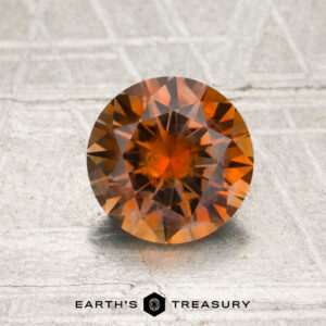 1.43-carat rich orange Montana sapphire (heated), round faceted, displayed on a textured light surface with the “EARTH’S TREASURY” logo and text at the bottom.