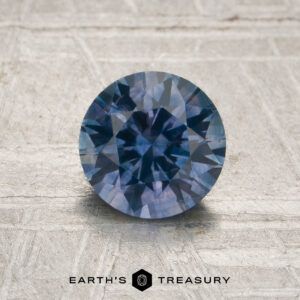 A 1.29-carat denim blue-purple particolored Montana sapphire (heated), with faceted cuts, is placed on a textured light surface, with the words “EARTH’S TREASURY” displayed below the stone.