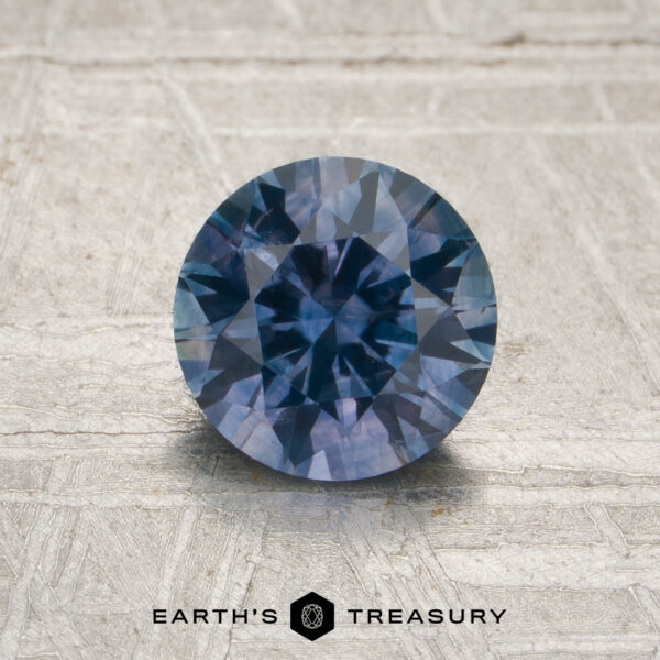 1.29-Carat Denim Blue-Purple Particolored Montana Sapphire (Heated) A 1.29-carat denim blue-purple particolored Montana sapphire (heated), with faceted cuts, is placed on a textured light surface, with the words “EARTH’S TREASURY” displayed below the stone.