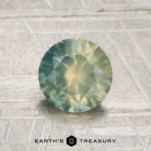 A round, faceted 1.25-carat opalescent Montana sapphire (heated) with green and yellow hues rests on a textured, light-colored surface. The “EARTH’S TREASURY” logo appears at the bottom of the image.