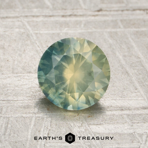 1.25-Carat Opalescent Light Aqua-Yellow Particolored Montana Sapphire (Heated) A round, faceted 1.25-carat opalescent Montana sapphire (heated) with green and yellow hues rests on a textured, light-colored surface. The “EARTH’S TREASURY” logo appears at the bottom of the image.