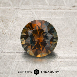 A 1.23-carat burnt orange-blue particolored Montana sapphire (heated), round and faceted, displaying vivid orange and brown hues, is set on a textured metallic surface with “EARTH’S TREASURY” branding at the bottom of the image.
