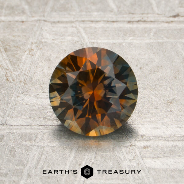 1.23-Carat Burnt Orange-Blue Particolored Montana Sapphire (Heated) A 1.23-carat burnt orange-blue particolored Montana sapphire (heated), round and faceted, displaying vivid orange and brown hues, is set on a textured metallic surface with “EARTH’S TREASURY” branding at the bottom of the image.