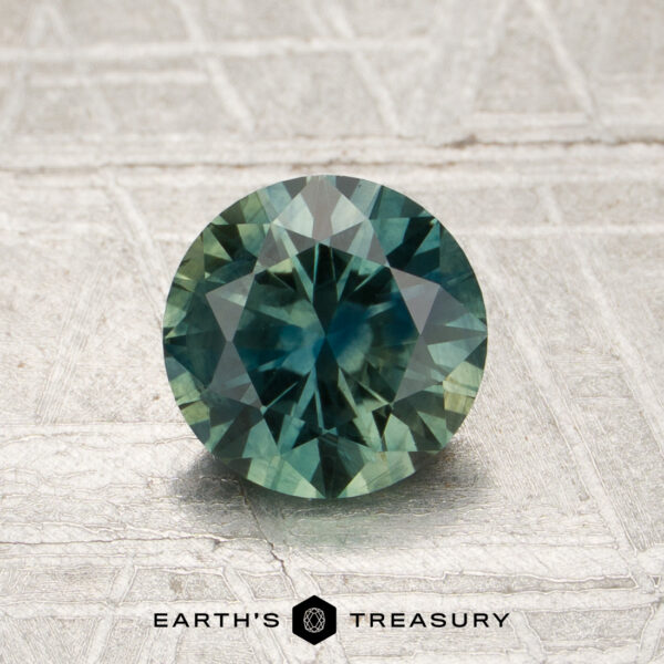 1.23-Carat Spruce Green Montana Sappire (Heated) A 1.23-carat spruce green Montana sapphire (heated), round-cut, showcased on a textured surface with the “Earth’s Treasury” text and hexagonal logo below the gemstone.