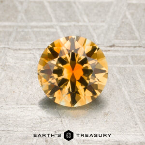 A round, faceted 1.21-carat golden yellow-orange particolored Montana sapphire is displayed on a metallic textured surface with the words “Earth’s Treasury” printed below.