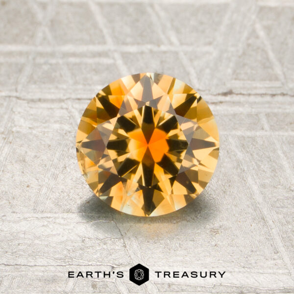 1.21-Carat Golden Yellow-Orange Particolored Montana Sapphire (Heated) A round, faceted 1.21-carat golden yellow-orange particolored Montana sapphire is displayed on a metallic textured surface with the words “Earth’s Treasury” printed below.