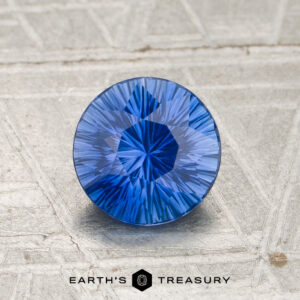 A round, faceted 1.03-carat rich blue Ceylon sapphire (heated) with an intricate cut is displayed upright on a textured light surface, with the “EARTH’S TREASURY” logo visible at the bottom.
