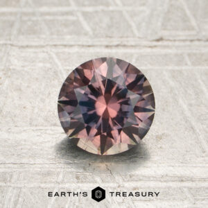 1.18-carat pink-purple particolored Montana sapphire (heated), round faceted, displayed on a textured surface above the Earth’s Treasury logo.