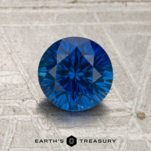 A 1.21-carat rich blue Madagascar sapphire (heated) with intricate faceting is displayed on a textured metallic surface, featuring the “EARTH’S TREASURY” logo and text at the bottom.