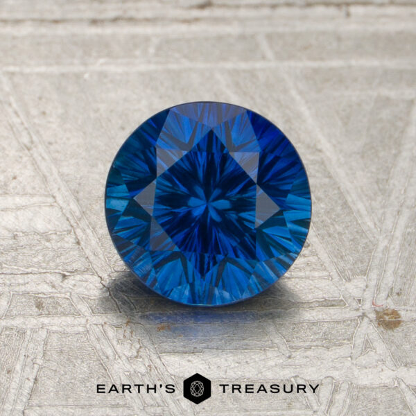 A 1.21-carat rich blue Madagascar sapphire (heated) with intricate faceting is displayed on a textured metallic surface, featuring the “EARTH’S TREASURY” logo and text at the bottom.