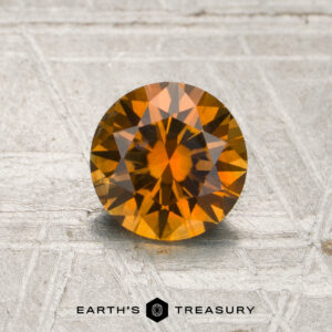 A round, faceted 1.07-carat rich orange Montana sapphire (heated) is displayed on a textured gray surface, with the text “EARTH’S TREASURY” and a hexagon logo beneath the stone.