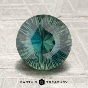 A round, teal-green 2.75-carat Madagascar sapphire with a faceted starburst pattern is displayed upright on a light textured surface. “Earth’s Treasury” is printed at the bottom of the image.