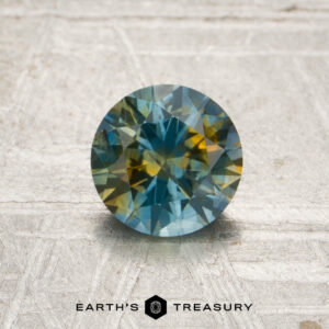 A 1.06-carat blue-yellow particolored Montana sapphire (heated), cut in a round shape, rests on a textured surface, with “Earth’s Treasury” and a geometric logo displayed below it.