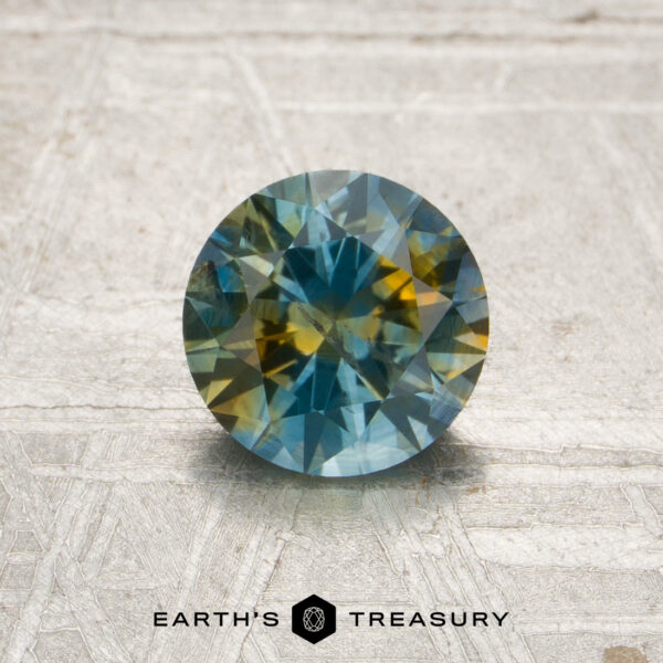 1.06-Carat Blue-Yellow Particolored Montana Sapphire (Heated) A 1.06-carat blue-yellow particolored Montana sapphire (heated), cut in a round shape, rests on a textured surface, with “Earth’s Treasury” and a geometric logo displayed below it.