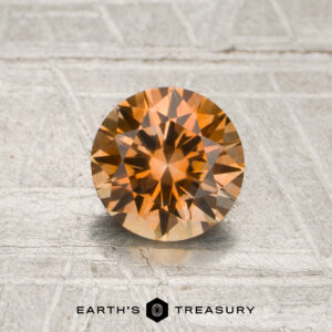 A round-cut 1.05-carat peachy orange Montana sapphire (heated) is displayed on a metallic surface, with the text “EARTH’S TREASURY” and a hexagonal logo at the bottom.