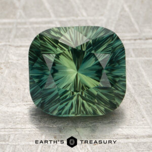 A 3.62-carat spruce green Madagascar sapphire (heated) with intricate faceting is displayed on a textured surface, accompanied by the text “EARTH’S TREASURY” and a geometric logo below.