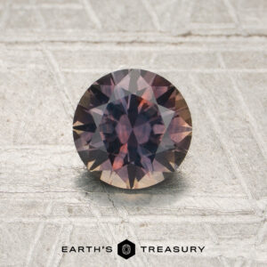 0.97-carat peach-purple particolored Montana sapphire (heated), round faceted, showcased on a textured surface with the “EARTH’S TREASURY” logo at the bottom.