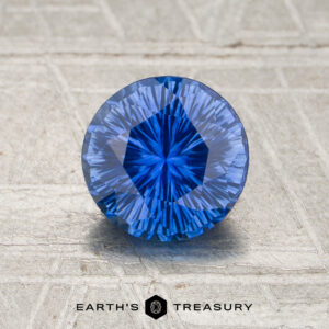 A 1.12-carat rich blue Ceylon sapphire (heated), cut in a round brilliant shape, sits on a textured surface with the words “EARTH’S TREASURY” and a hexagonal logo beneath it.