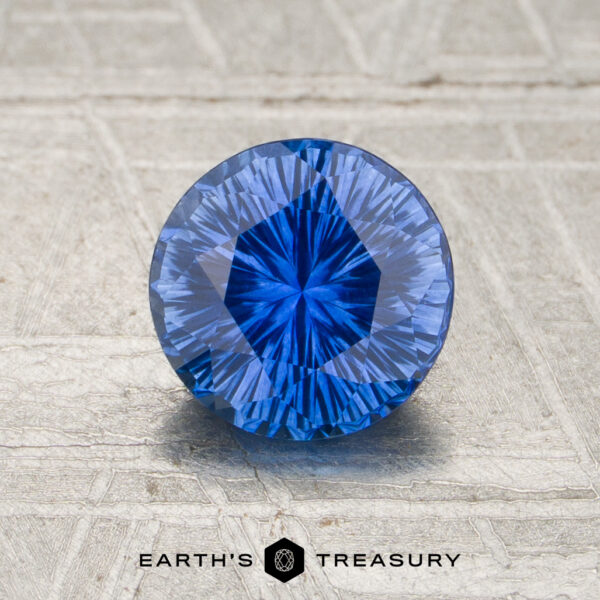 1.12-Carat Rich Cornflower Blue Ceylon Sapphire (Heated) A 1.12-carat rich blue Ceylon sapphire (heated), cut in a round brilliant shape, sits on a textured surface with the words “EARTH’S TREASURY” and a hexagonal logo beneath it.