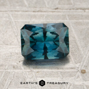 A rectangular step-cut 1.45-carat peacock blue Montana sapphire (heated) is placed on a textured surface with the words “EARTH’S TREASURY” and a hexagonal logo below it.