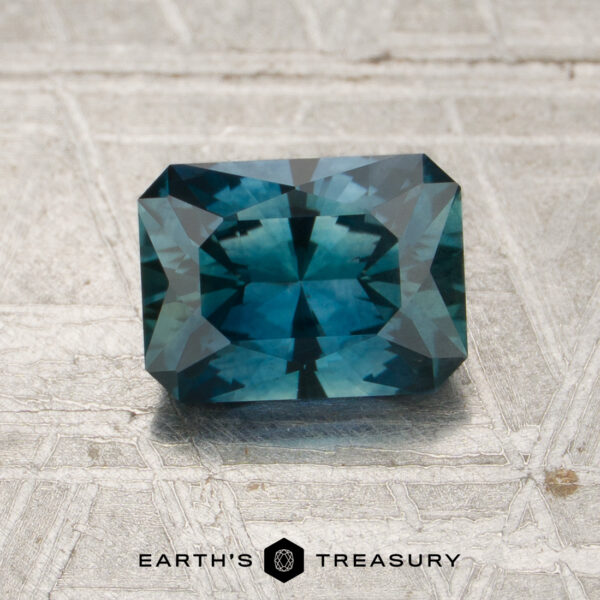 1.45-Carat Peacock Blue Montana Sapphire (Heated) A rectangular step-cut 1.45-carat peacock blue Montana sapphire (heated) is placed on a textured surface with the words “EARTH’S TREASURY” and a hexagonal logo below it.