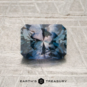 A rectangular, blue-pink particolored faceted Montana sapphire (1.32 carats, heated) rests on a metallic textured surface. The logo “EARTH’S TREASURY” appears at the bottom of the image.