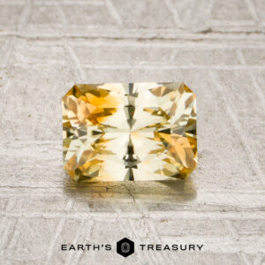 A 1.21-carat pale yellow-orange particolored Montana sapphire with radiant cut facets is displayed on a textured gray surface, with the “EARTH’S TREASURY” logo visible at the bottom of the image.